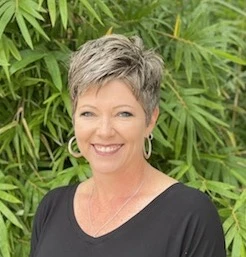 Woman with short blonde hair wearing a black v-neck top, smiling in front of green foliage.