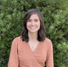 Woman with dark hair wearing peach-colored V-neck top smiling at camera against green foliage background