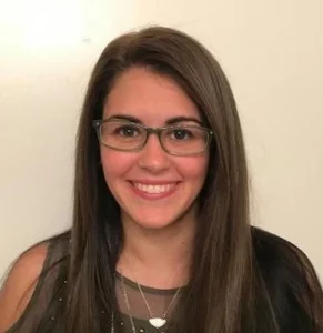 Young woman with long dark hair and glasses wearing dark clothing, smiling at camera against a white background.