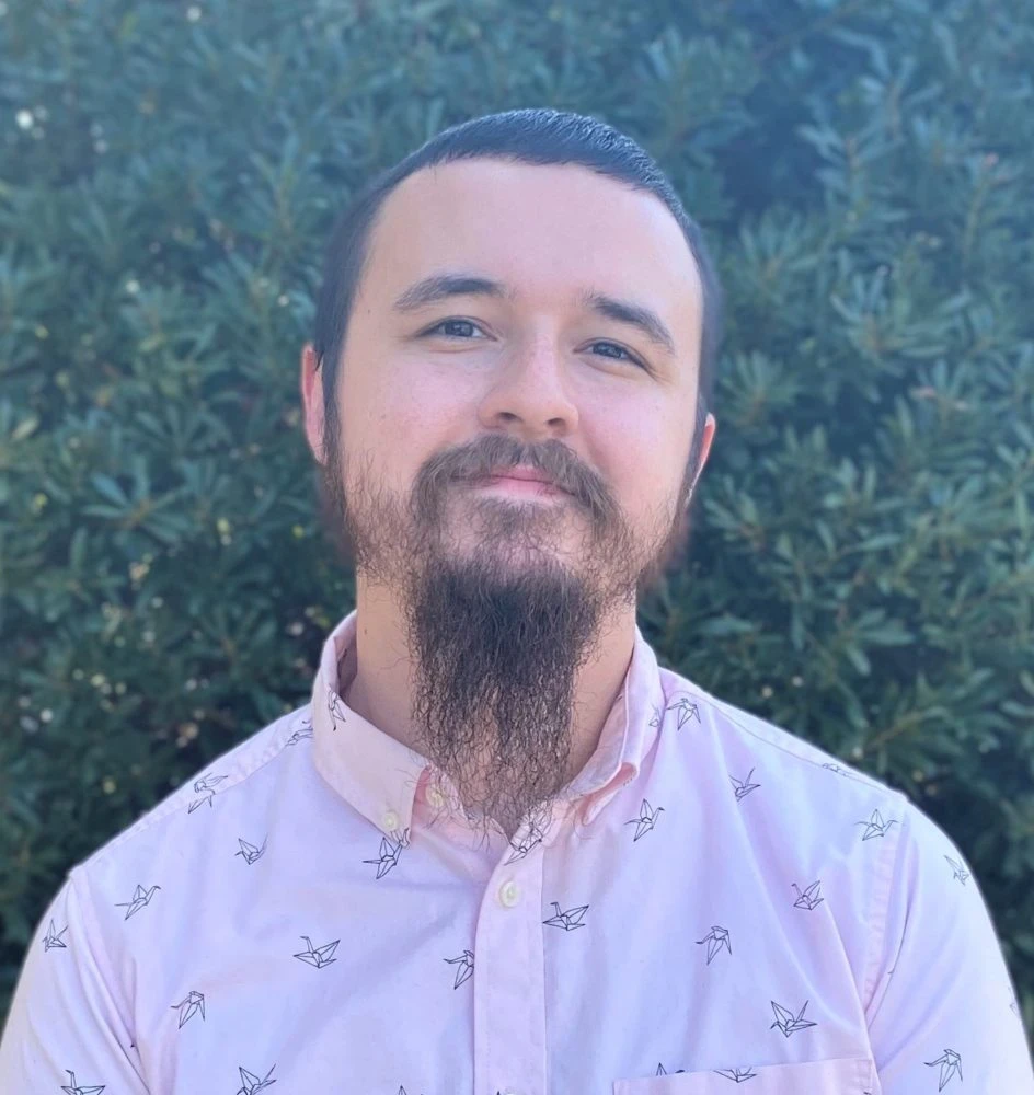 Man with dark hair and a full beard wearing a light pink button-up shirt with a small pattern, photographed against green foliage.