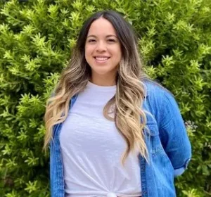 Young woman with long brown and blonde ombre hair smiling at camera, wearing light blue t-shirt and blue cardigan, green foliage