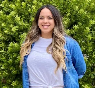 Young woman with long brown and blonde ombre hair smiling at camera, wearing light blue t-shirt and blue cardigan, green foliage