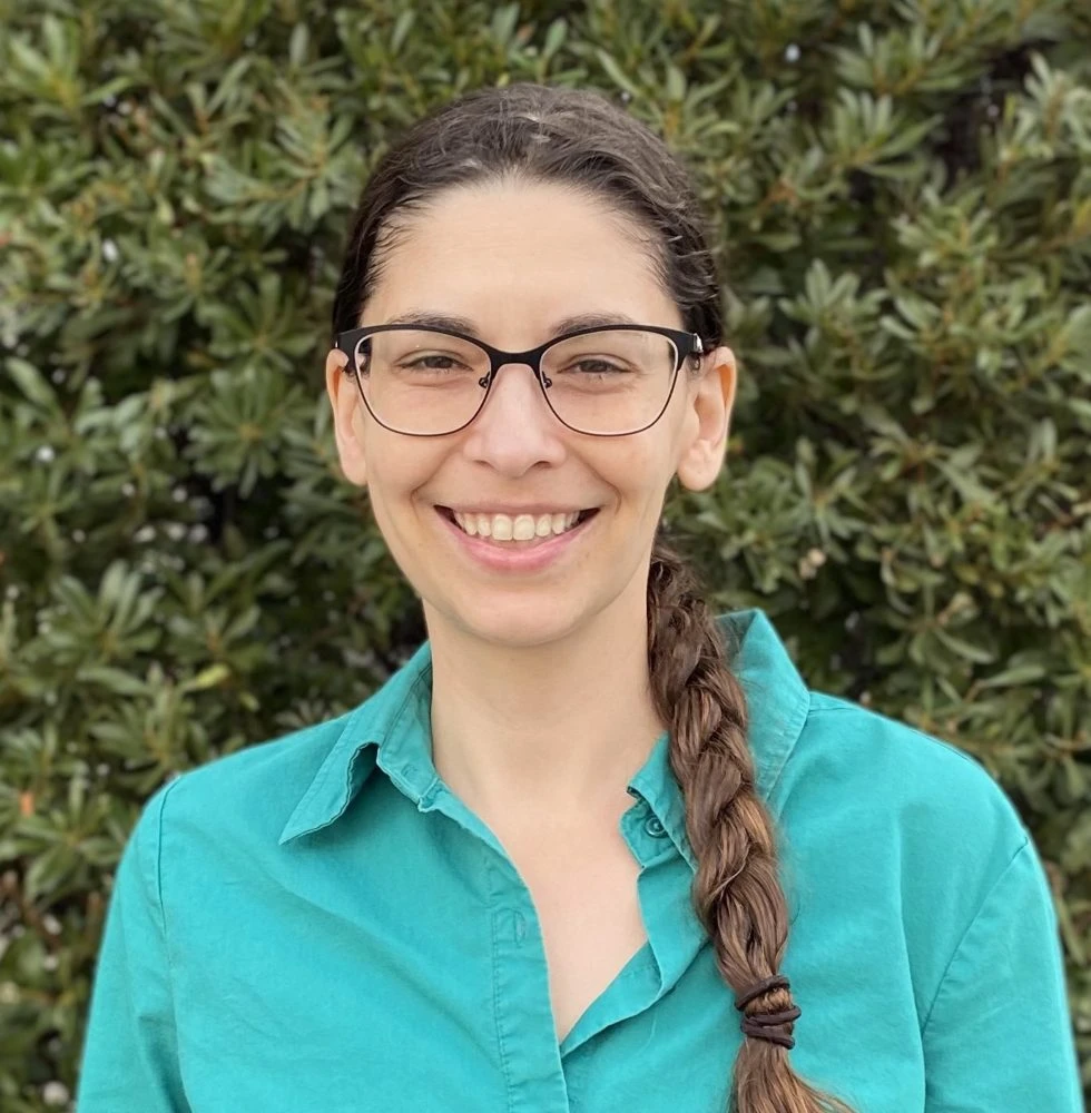 Woman with brown braided hair wearing teal button-up shirt smiling at camera against green foliage