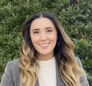Young woman with long brown wavy hair smiling outdoors against green foliage, wearing a gray cardigan over a cream colored sweater.