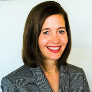 Woman with shoulder-length dark hair and red lipstick, wearing a checkered gray blazer, smiling at camera against white background