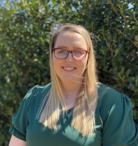 Woman with long blonde hair, glasses, and a green shirt smiling outdoors with green foliage background