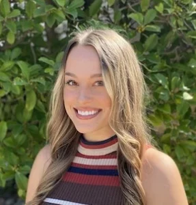 Woman with long blonde hair smiling at camera, wearing striped sleeveless tank top in pink and burgundy, green foliage background