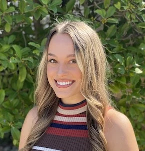 Woman with long blonde hair smiling at camera, wearing striped sleeveless tank top in pink and burgundy, green foliage background