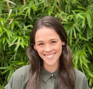 Woman with long dark hair smiling at camera, wearing olive green collared shirt against green foliage background