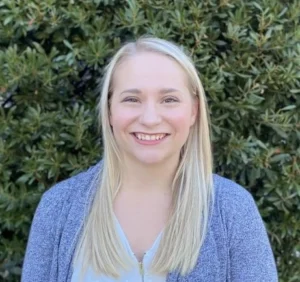 Young woman with long blonde hair smiling at camera wearing gray cardigan against green foliage background
