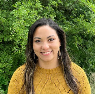 Young woman with long dark hair wearing a mustard yellow knit sweater, smiling outdoors with green foliage in the background.