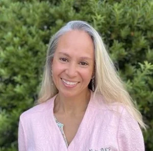 Smiling woman with long blonde hair wearing light pink cardigan photographed against blurred green hedge background.
