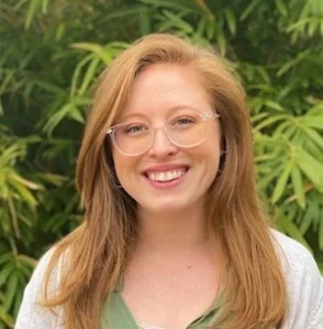 Woman with reddish-blonde hair wearing glasses and a white top, smiling at the camera outdoors against green foliage background.