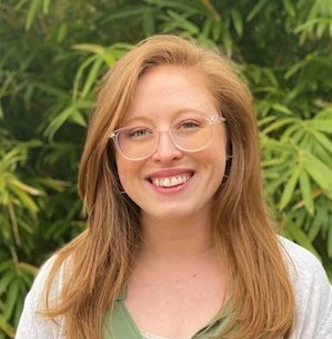 Woman with reddish-blonde hair wearing glasses and a white top, smiling at the camera outdoors against green foliage background.