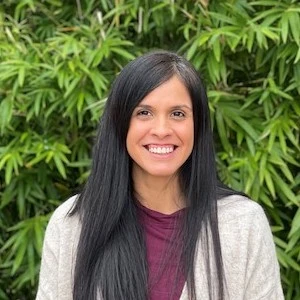 Woman with long black hair and purple top smiling at camera with green foliage background