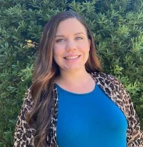 Woman with long dark hair wearing a blue top and leopard print cardigan, smiling outdoors with green background
