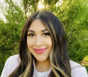 Woman with long dark hair with blonde highlights, wearing a white top, smiling outdoors with green foliage