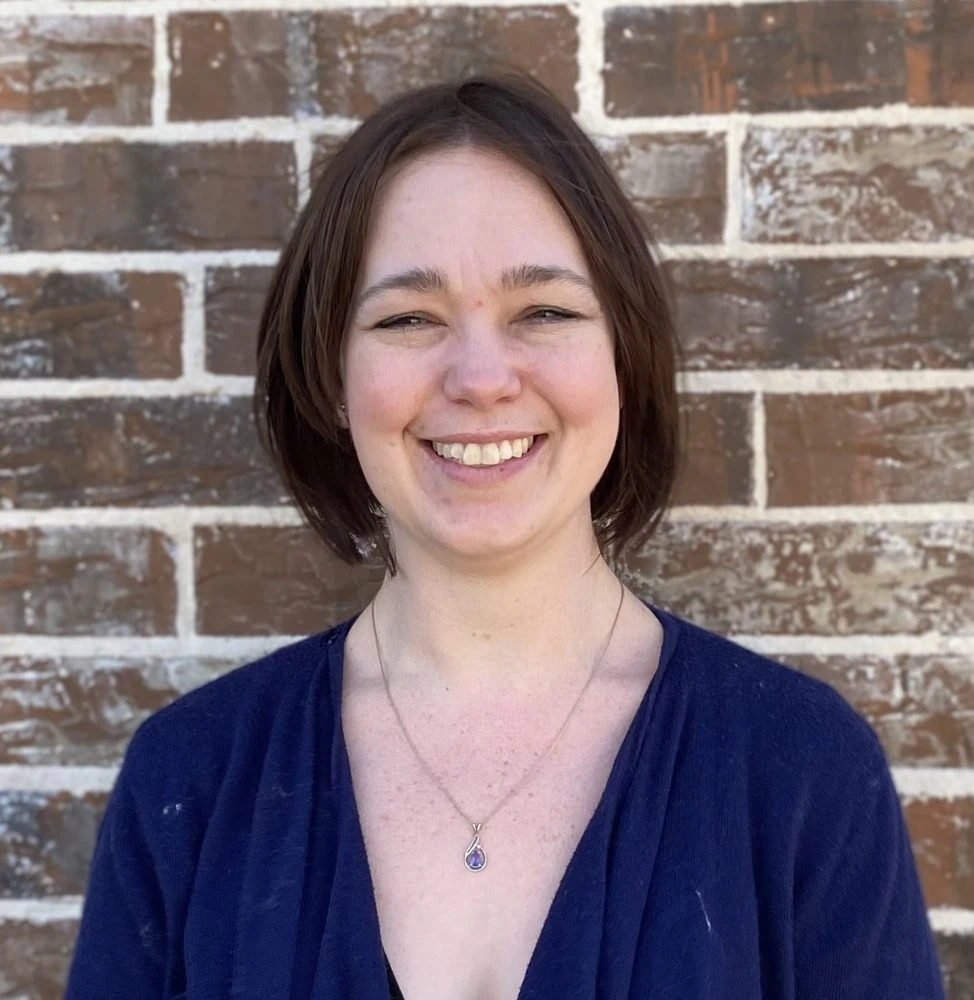 Woman with dark shoulder-length hair smiling at camera, wearing dark blue v-neck shirt with pendant necklace, brick wall background