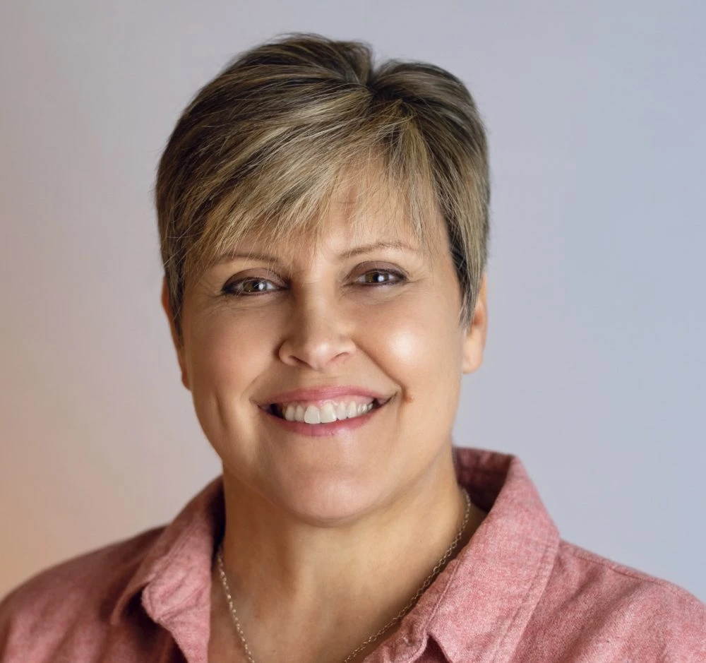 Woman with short blonde and brown hair wearing pink shirt, smiling at camera against neutral background