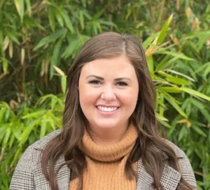 Woman with long brown hair wearing a mustard turtleneck and plaid blazer, smiling outdoors