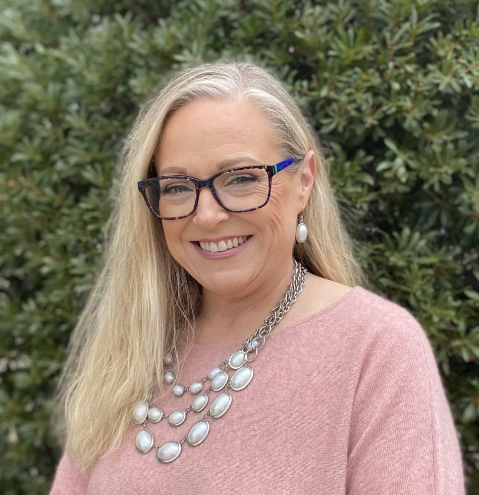 Woman with blonde hair and blue-framed glasses wearing pink sweater with pearl necklace smiling