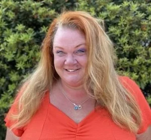Smiling woman with long reddish-blonde hair wearing bright red v-neck top photographed outdoors against blurred green background.