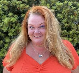 Smiling woman with long reddish-blonde hair wearing bright red v-neck top photographed outdoors against blurred green background.