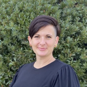 Woman with short dark hair wearing black shirt, smiling at camera against green foliage background