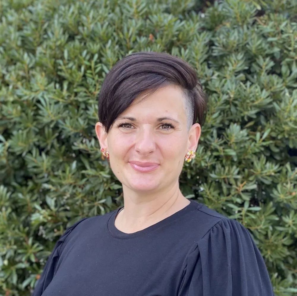 Woman with short dark hair wearing black shirt, smiling at camera against green foliage background