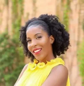 Smiling woman with dark curly hair wearing bright yellow sleeveless top photographed outdoors against blurred green foliage background.