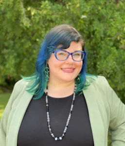 Woman with teal and dark hair, glasses, wearing a light green cardigan and beaded necklace with green foliage