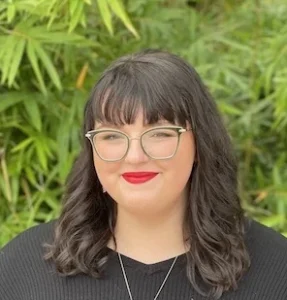 Woman with dark wavy hair and bangs wearing glasses and red lipstick, smiling at camera with green plant background