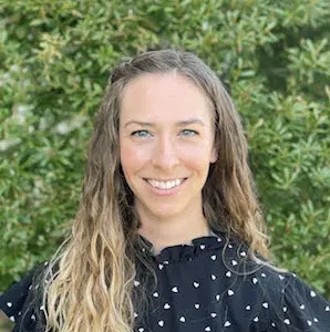 Woman with long blonde wavy hair wearing a black top with white dotted pattern, smiling at camera outdoors with green foliage background.