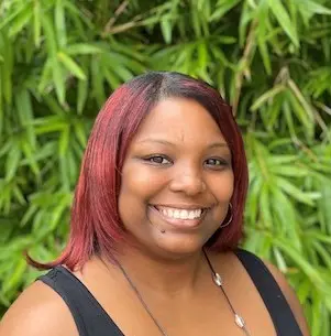 Woman with pink-tinted dark hair wearing a black sleeveless top, smiling at camera with green foliage visible in background.
