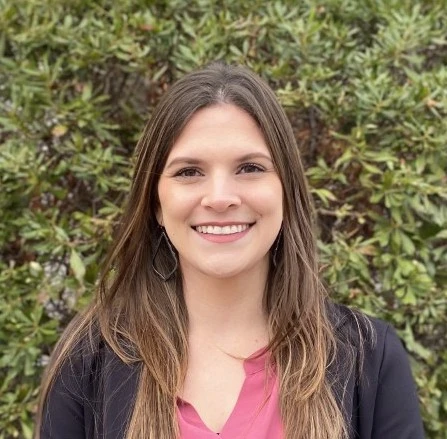 Young woman with long dark hair with highlights wearing pink top and black cardigan, smiling outdoors