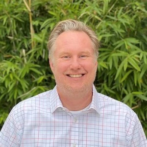 Man with light colored hair wearing a light blue and pink checkered button-up shirt, smiling outdoors against green foliage background.