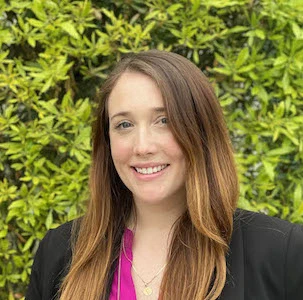 Woman with long reddish-brown hair smiling at camera, wearing black blazer over pink top, green foliage background