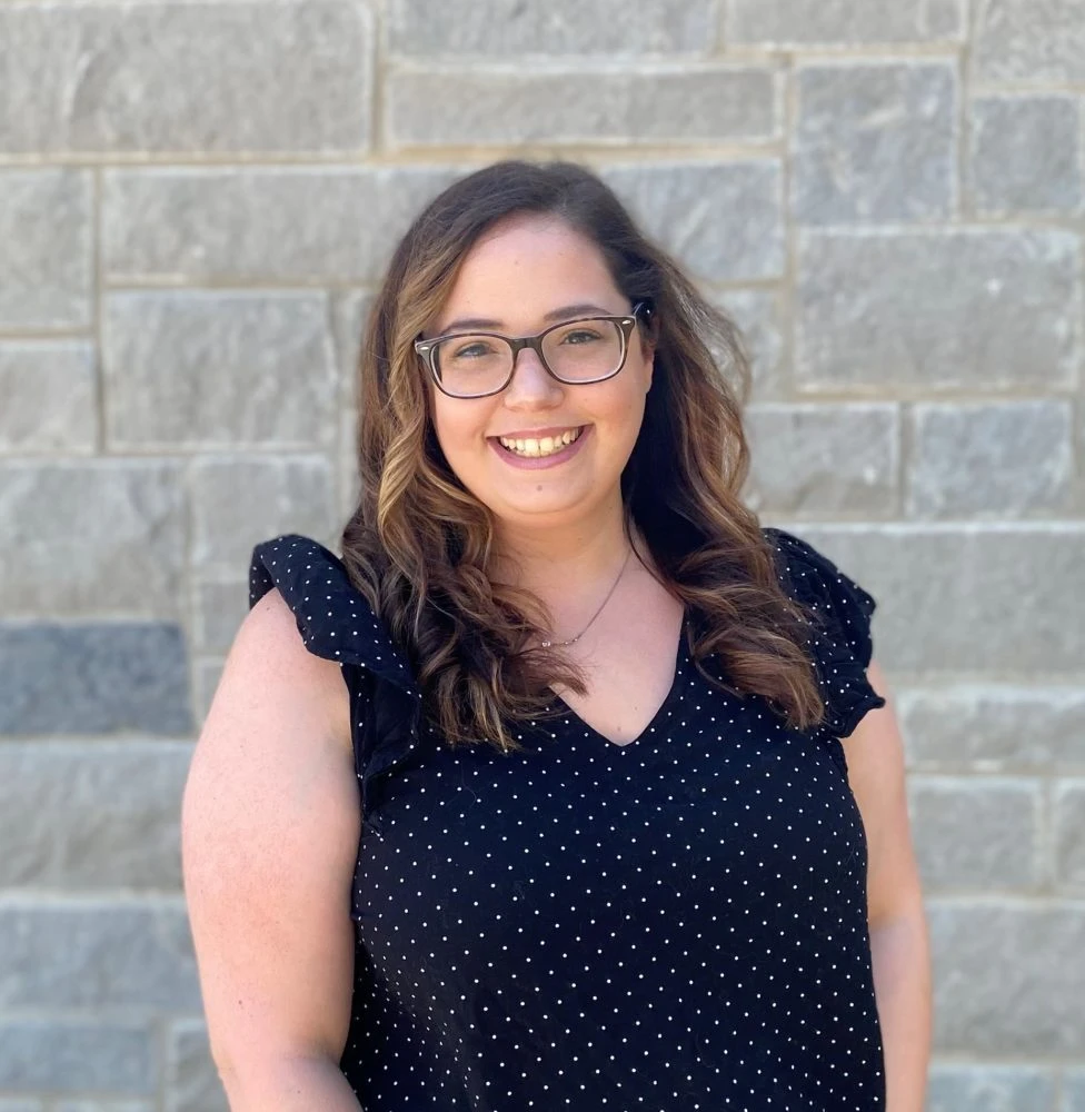 Woman with brown hair wearing glasses and black polka-dot top smiling at camera against tan brick wall