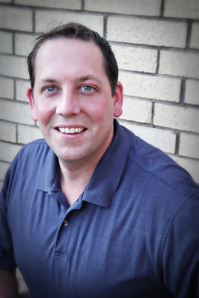 Man with short dark hair wearing a dark blue polo shirt smiling against a brick wall background