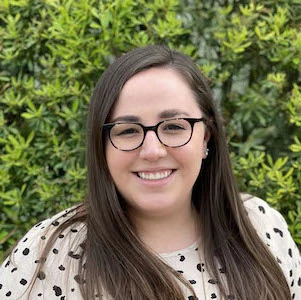 Woman with long dark hair and glasses wearing a polka dot top, smiling at the camera against a green background
