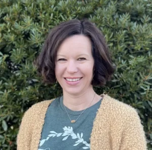 Woman with shoulder-length dark hair wearing a teal shirt and tan cardigan, smiling at the camera with green foliage in the background.