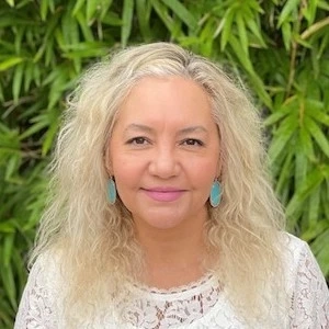 Woman with long blonde wavy hair wearing turquoise earrings and white lace top, smiling against green foliage background