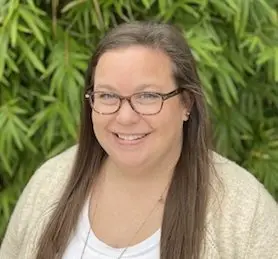 Young woman with long dark hair and glasses wearing white top, smiling outdoors with greenery background