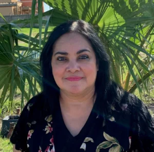 A woman with long dark hair wearing pink lipstick and a black floral patterned top outdoors with green foliage