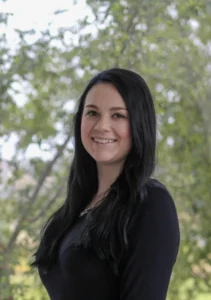 Woman with long dark hair wearing a black top, smiling at camera with trees visible in background