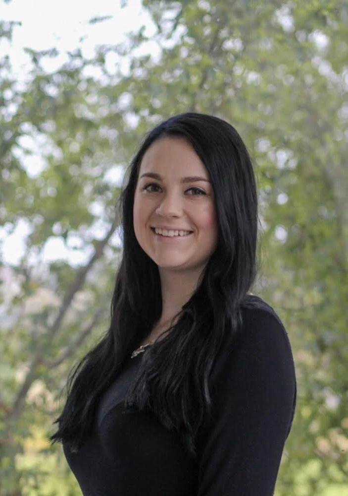 Woman with long dark hair wearing a black top, smiling at camera with trees visible in background