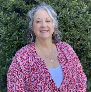 Woman with gray and white hair wearing pink patterned cardigan over blue top, smiling outdoors against green foliage