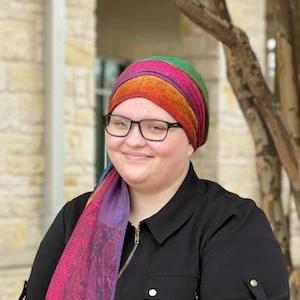 Portrait of a smiling person wearing glasses and a colorful rainbow striped headwrap with a black shirt against an indoor background