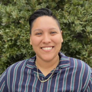 Person with dark slicked-back hair wearing navy striped shirt with gold chain necklace, smiling at camera with green plants