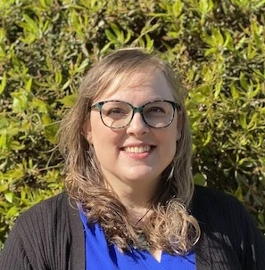 Woman with long dark hair and glasses wearing a dark blazer over blue top, smiling outdoors with green foliage
